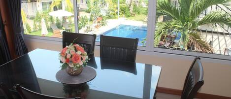 Un comedor con mesa y sillas de vidrio, un jarrón de flores y vistas a una piscina y edificios.