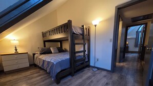 Habitación con literas, un escritorio y lámpara, suelo de madera y una puerta que da a otra habitación.