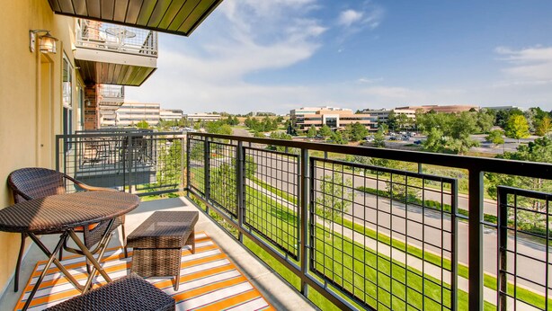Ein Balkon mit Korbmöbeln, einer Streifenoptik-Bereitung und Blick auf einen Parkplatz sowie Gebäude.