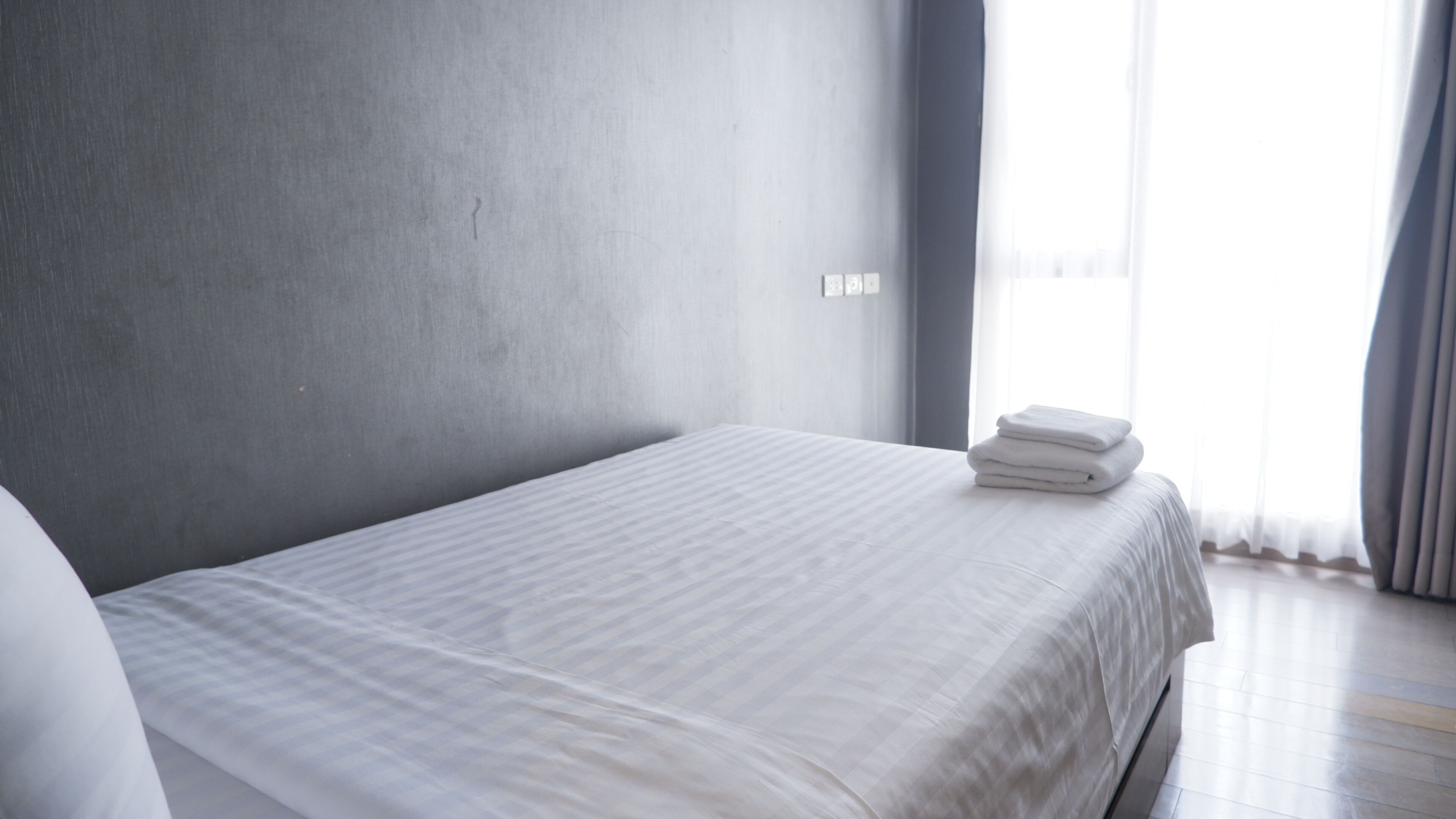 A minimalist hotel room with a large bed, a grey wall, and a window with curtains.