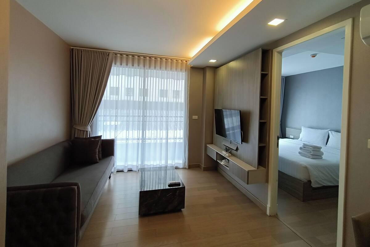 Una habitación de hotel moderna con sofá, televisor, cama y un ventanal con cortinas.