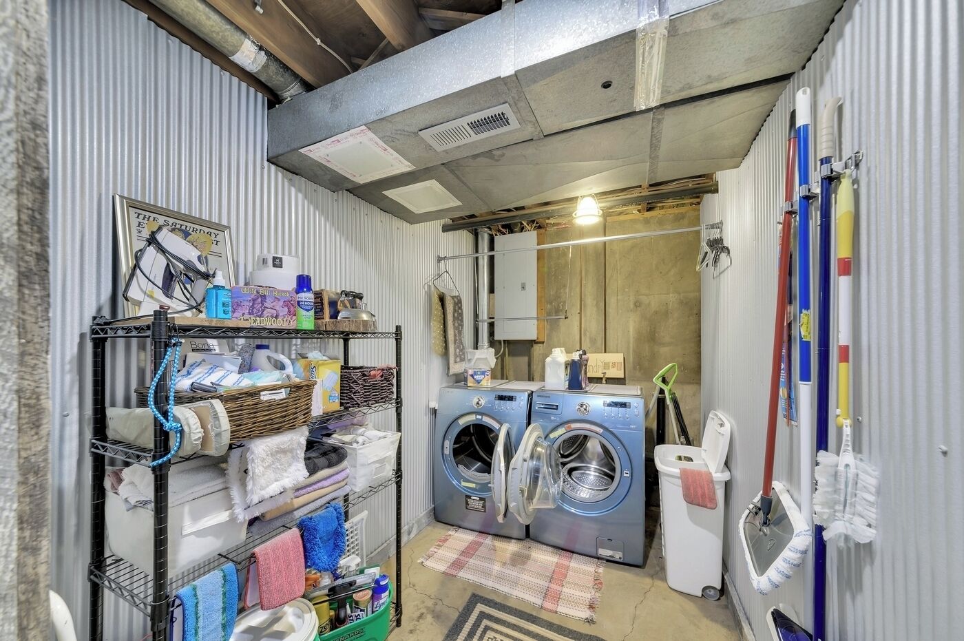 A laundry room with two front-loading washing machines, a metal shelving unit with various cleaning supplies, and a small rug on the floor.