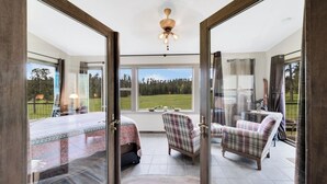 Una habitación amplia con una cama grande, dos sillones a cuadros, un escritorio con lámpara y vistas a un paisaje verde a través de amplios ventanales.