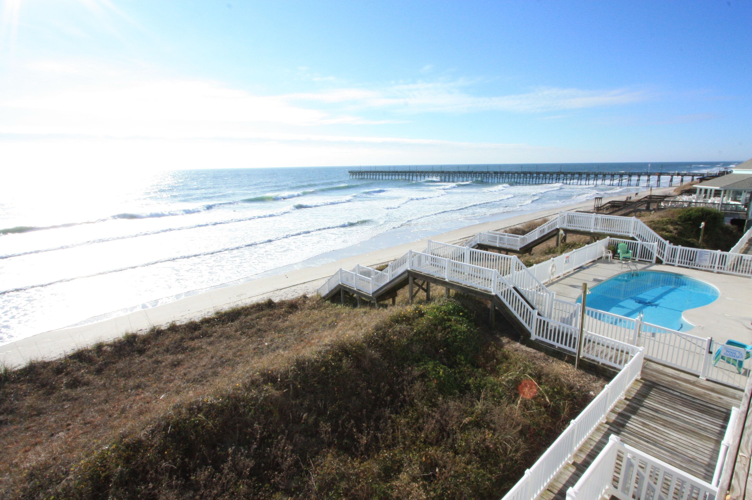 Une vue sur la plage avec une jetée qui s’avance dans l’océan, une piscine et une clôture blanche qui entoure le site.