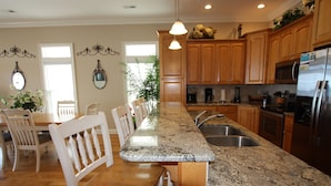 A kitchen with wooden cabinets, granite countertops, a stainless steel refrigerator, and a dining area with white chairs and a wooden table.