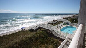 A beach view with a pier, a swimming pool, and a deck area with chairs and umbrellas.