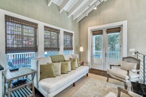 Un salon confortable avec un canapé, un fauteuil et une petite table sur laquelle se trouve un appareil. De grandes fenêtres avec des stores et une porte donnant sur un balcon sont visibles.