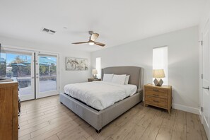 Ein Schlafzimmer mit einem Bett, einem Deckenventilator, einem Nachttisch, einer Lampe und Blick durch eine Glastür auf einen Außenbereich.
