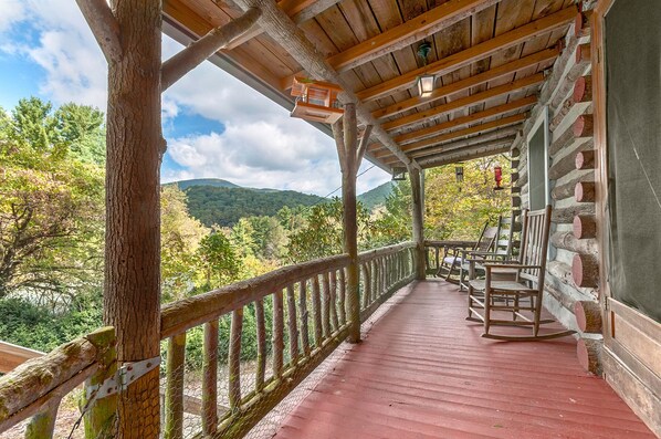 Una terraza de madera con piso rojo, rodeada por barandilla de madera, con vista a una zona boscosa y una montaña al fondo.