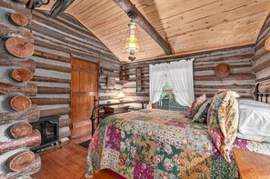 Un dormitorio de cabaña de troncos con una cama, una puerta de madera, un espejo y una ventana con cortina blanca.