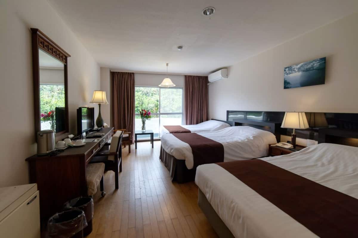 Ein Hotelzimmer mit zwei Betten, einem Schreibtisch, einem Stuhl, einem Spiegel, einem Fenster mit Vorhängen und einer Wandklimaanlage.