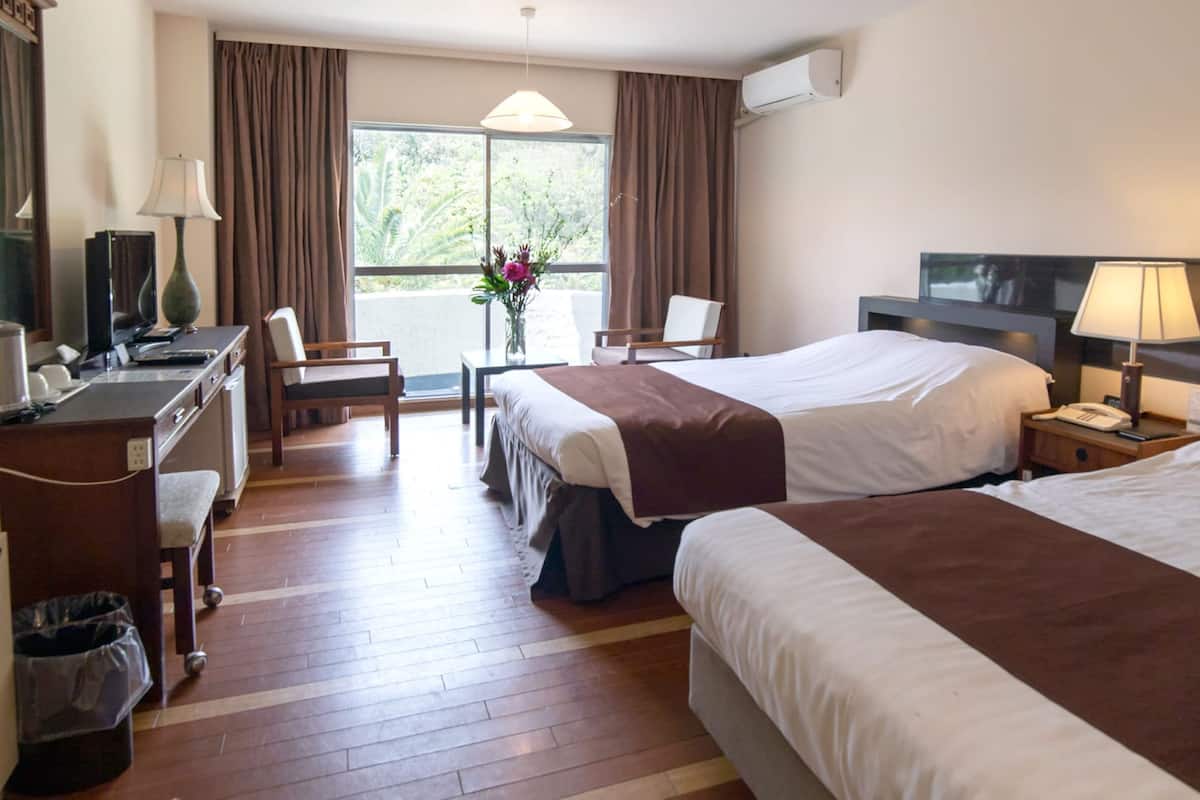 Ein Hotelzimmer mit zwei Betten, einem Schreibtisch, einem Stuhl, einem kleinen Tisch mit einer Vase und einem Fenster mit Vorhängen.