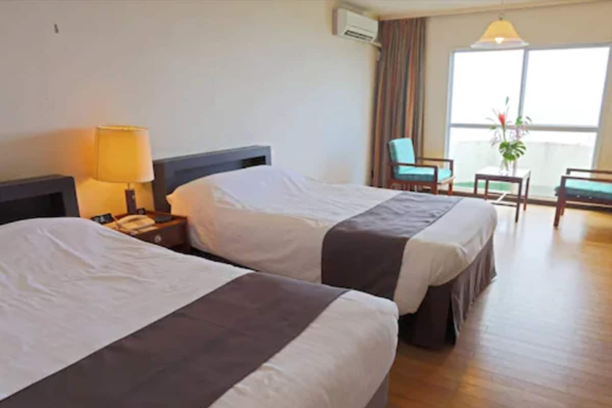 Ein Hotelzimmer mit zwei Betten, einem Schreibtisch mit Lampe, einem Sessel und einem Fenster mit Vorhängen.