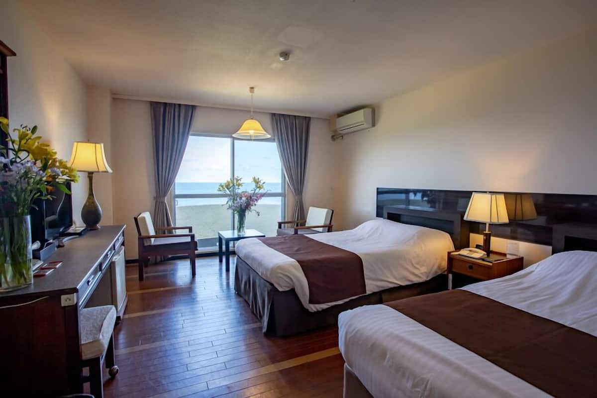 Ein Hotelzimmer mit zwei Betten, einem Schreibtisch, einem Stuhl, einer Lampe und Meerblick.