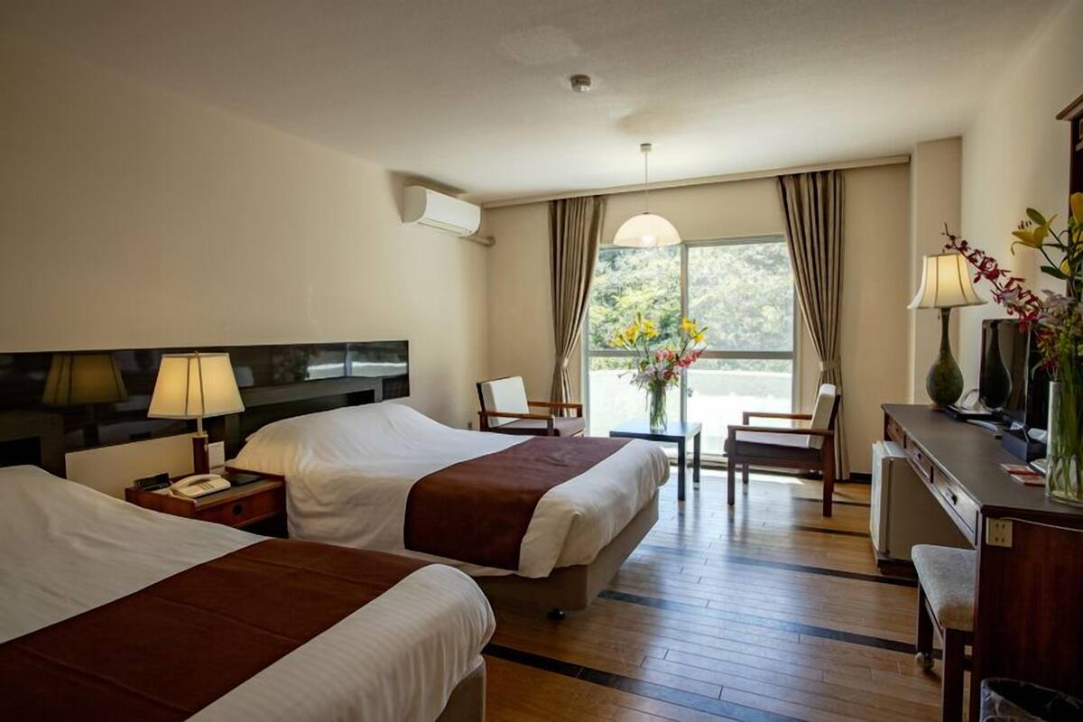 Ein Hotelzimmer mit zwei Betten, einem Schreibtisch, einem Stuhl, einer Lampe und einem Fenster mit Vorhängen.