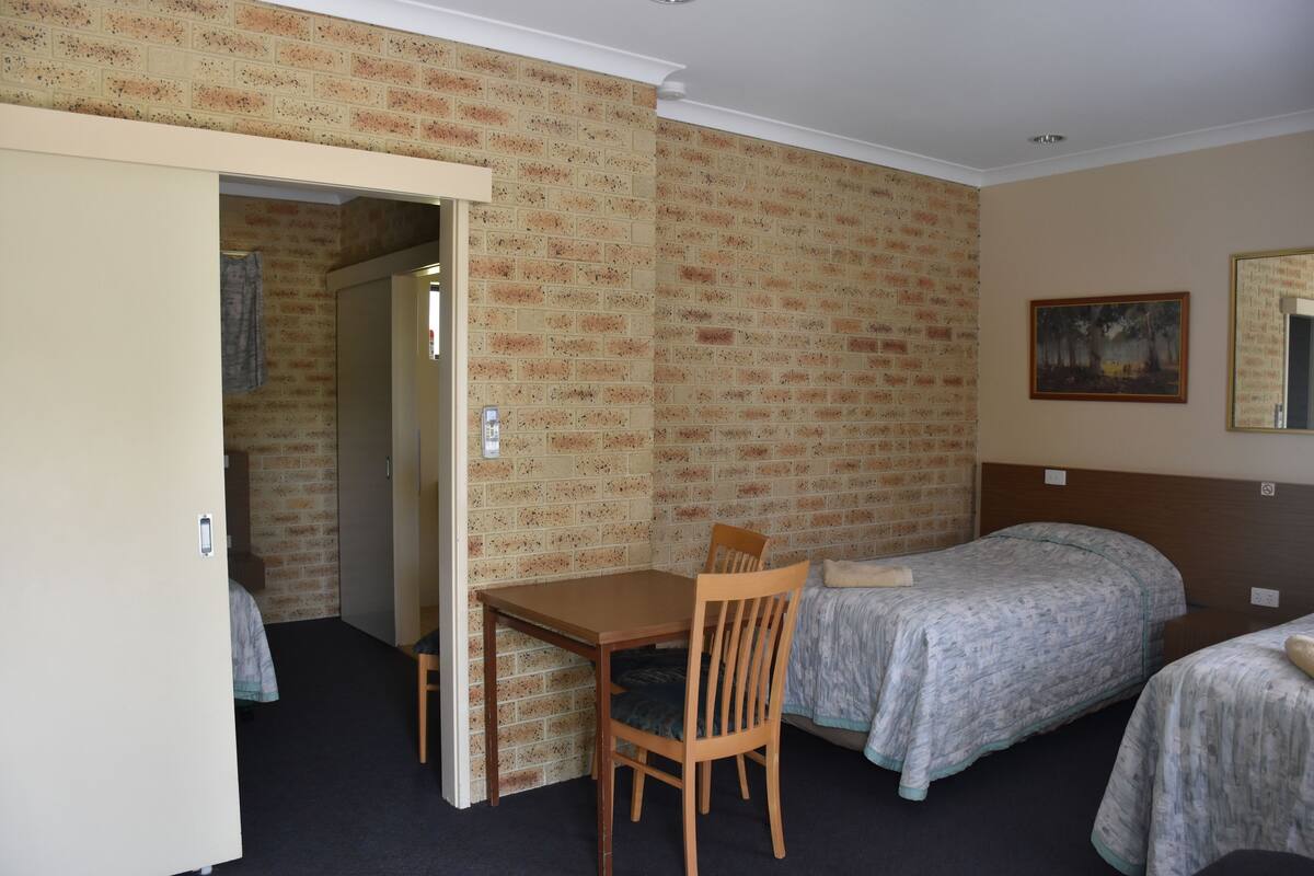 A room with a brick-patterned wall, a wooden table with a chair, two beds with grey bedding, and a painting on the wall.