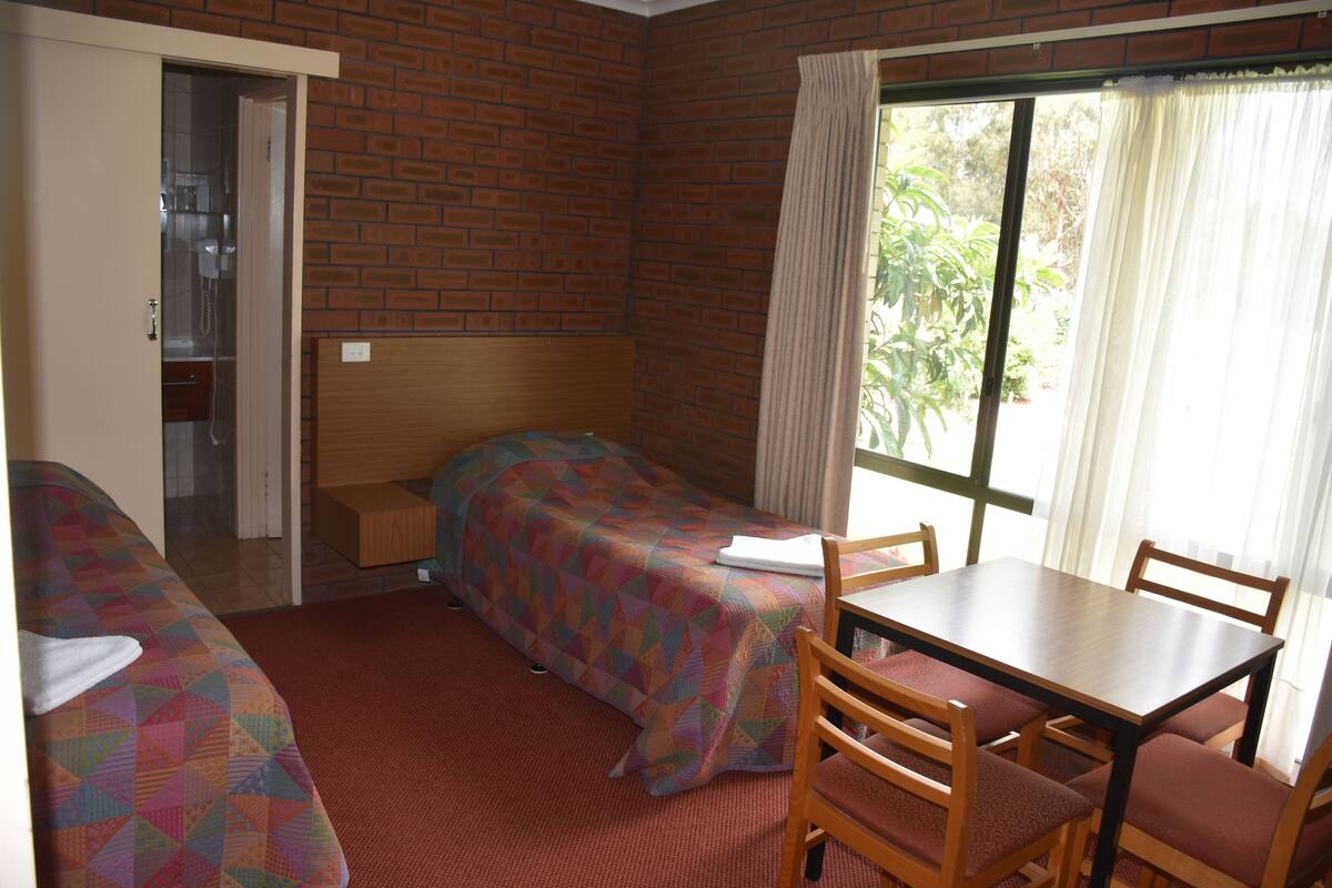 A room with two beds, a wooden table, and chairs. There is a window with curtains, and a door leading to a bathroom.