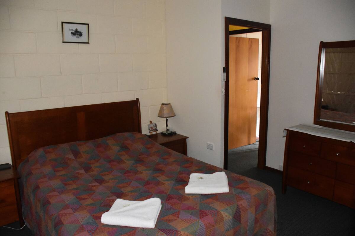 A hotel room with a wooden bed, a nightstand with a lamp, a dresser, and a framed picture on the wall.