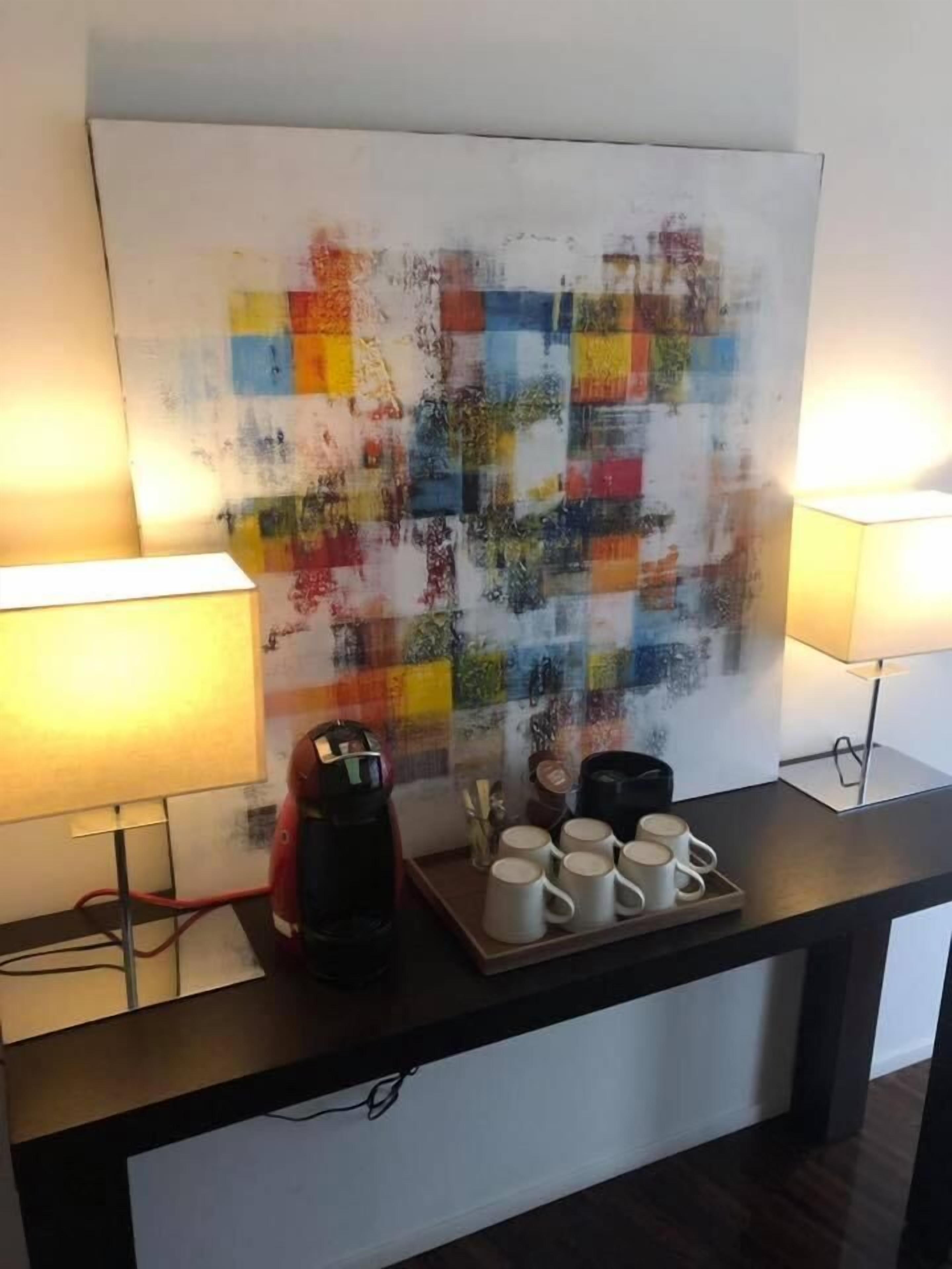 A coffee station with a coffee maker, mugs, and a colorful abstract painting on the wall.