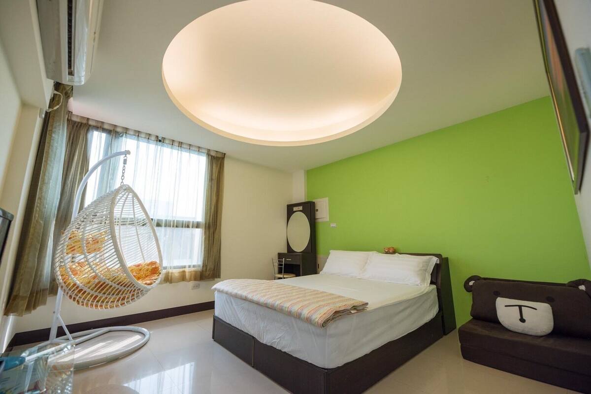 A hotel room with a bed, a hanging chair, a green wall, and a ceiling light.