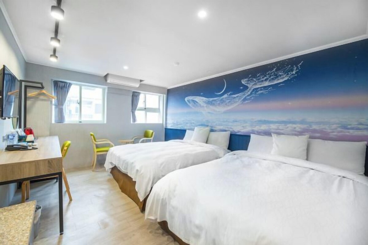 A hotel room with two beds, a desk, and a chair. There is a large window and a wall mural of space.