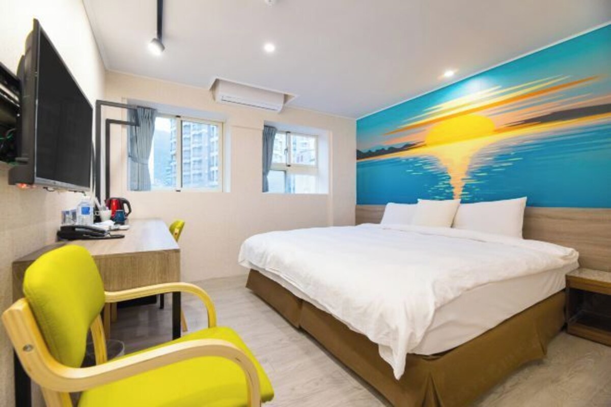 Habitación de hotel con cama, escritorio, silla y una pintura de un atardecer sobre el agua.