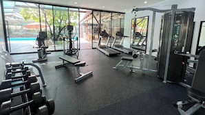 A modern gym with cardio equipment, weight machines, and a large window offering a view of the outdoors. - Glai Gan Place Hotel (Saraburi)