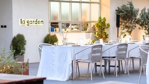 An outdoor dining area with a table set for four, surrounded by wicker chairs, in front of a building with a large window and the text "la garden" on the wall. - Glai Gan Place Hotel (Saraburi)