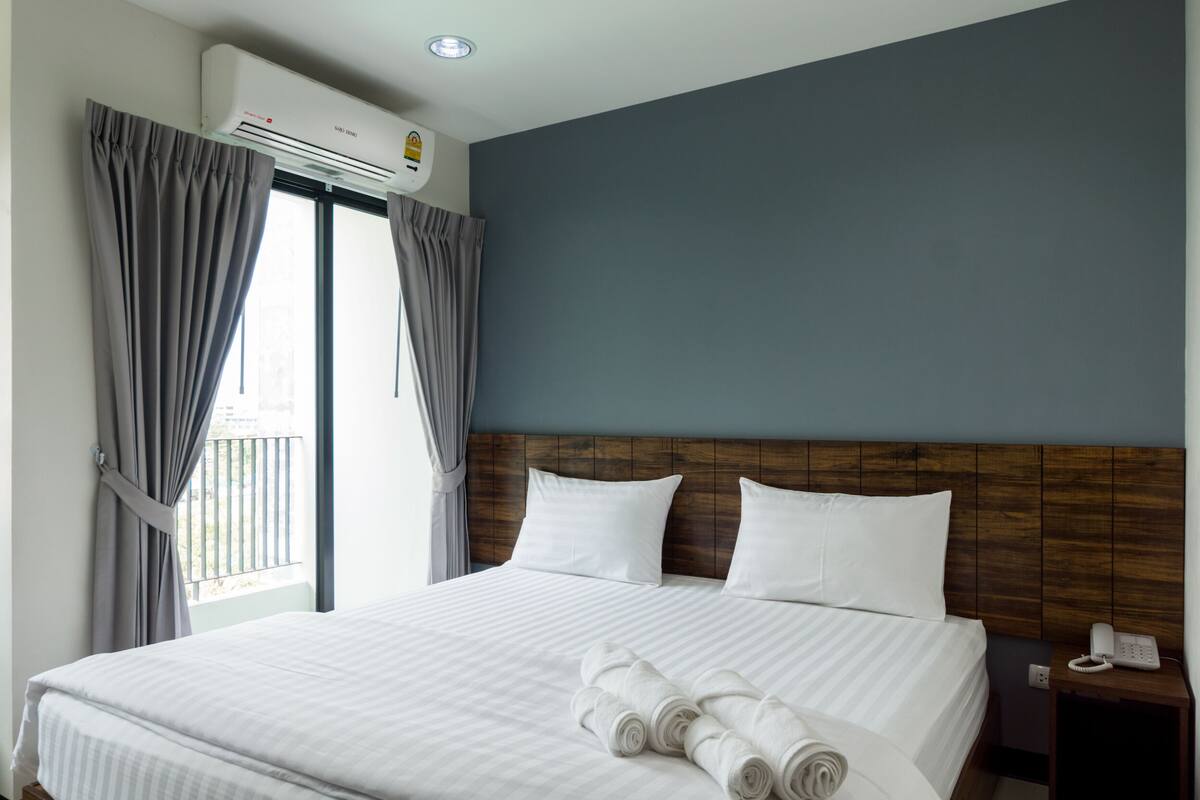 Una habitación de hotel con una cama grande, cortinas grises, un cabecero de madera y un aire acondicionado montado en la pared.