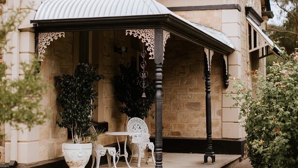 A stone building with a covered porch, a chimney, and a small garden area with a table and chairs. - Azalea Cedar-Rose Villa (Lyndoch)