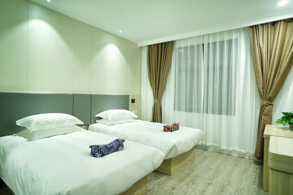 Ein Hotelzimmer mit zwei Betten, einem Holzkopfende, einem Nachttisch mit Pflanze und transparenten Vorhängen.
