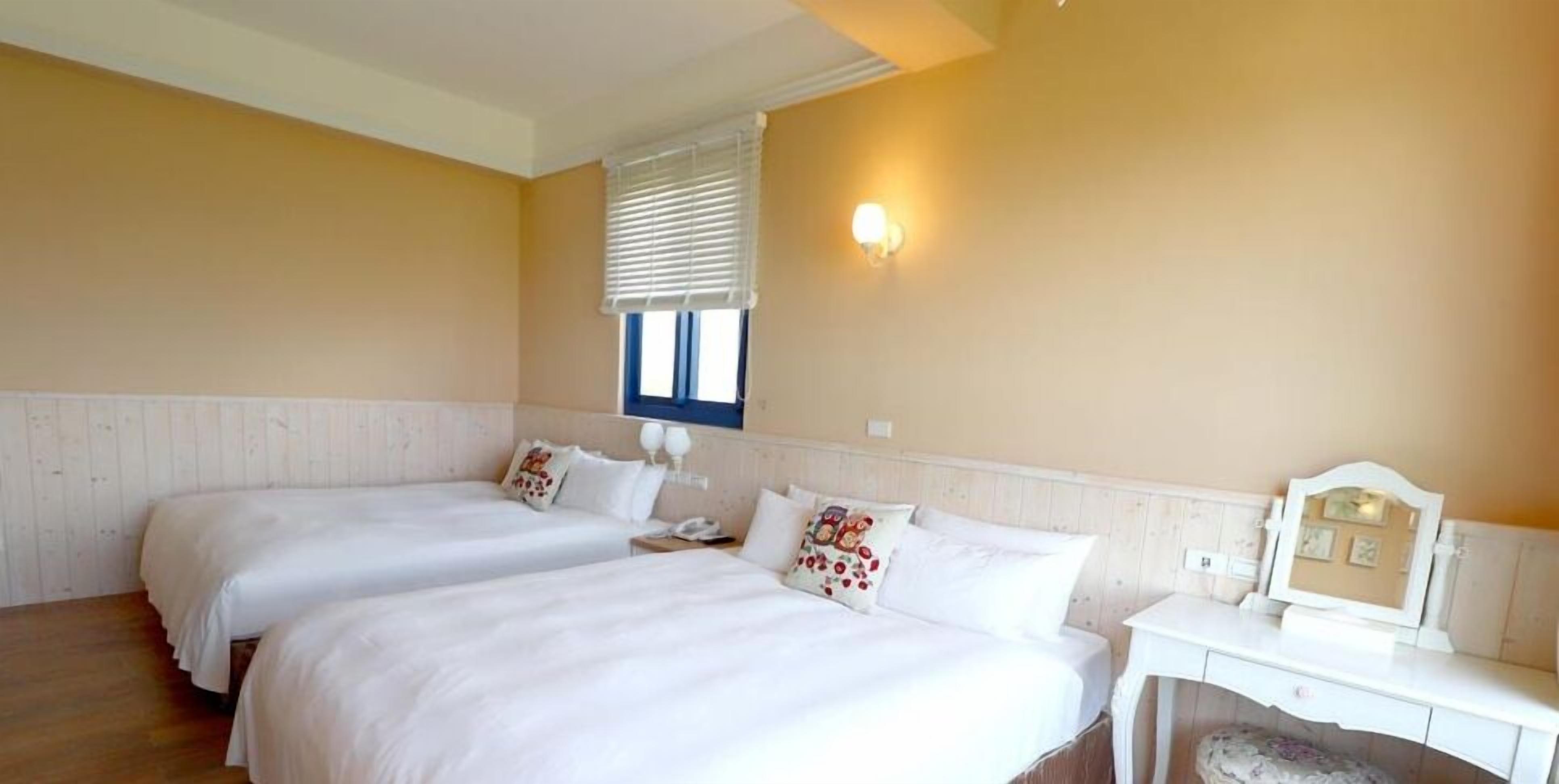 A hotel room with two beds, a bedside table with a lamp, a mirror, and a window with blinds.