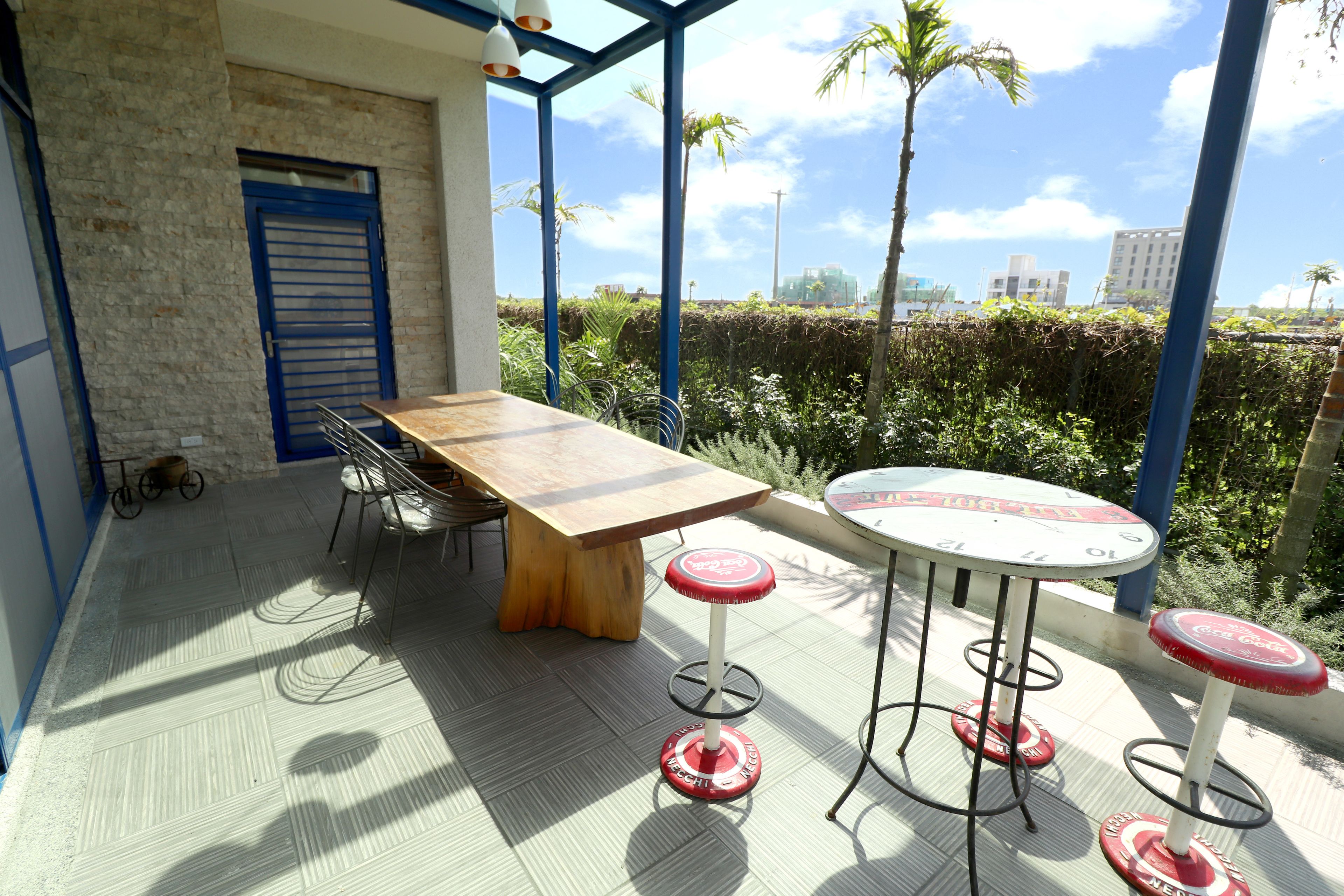 An outdoor patio with a wooden table and red bar stools, surrounded by greenery and a view of buildings.