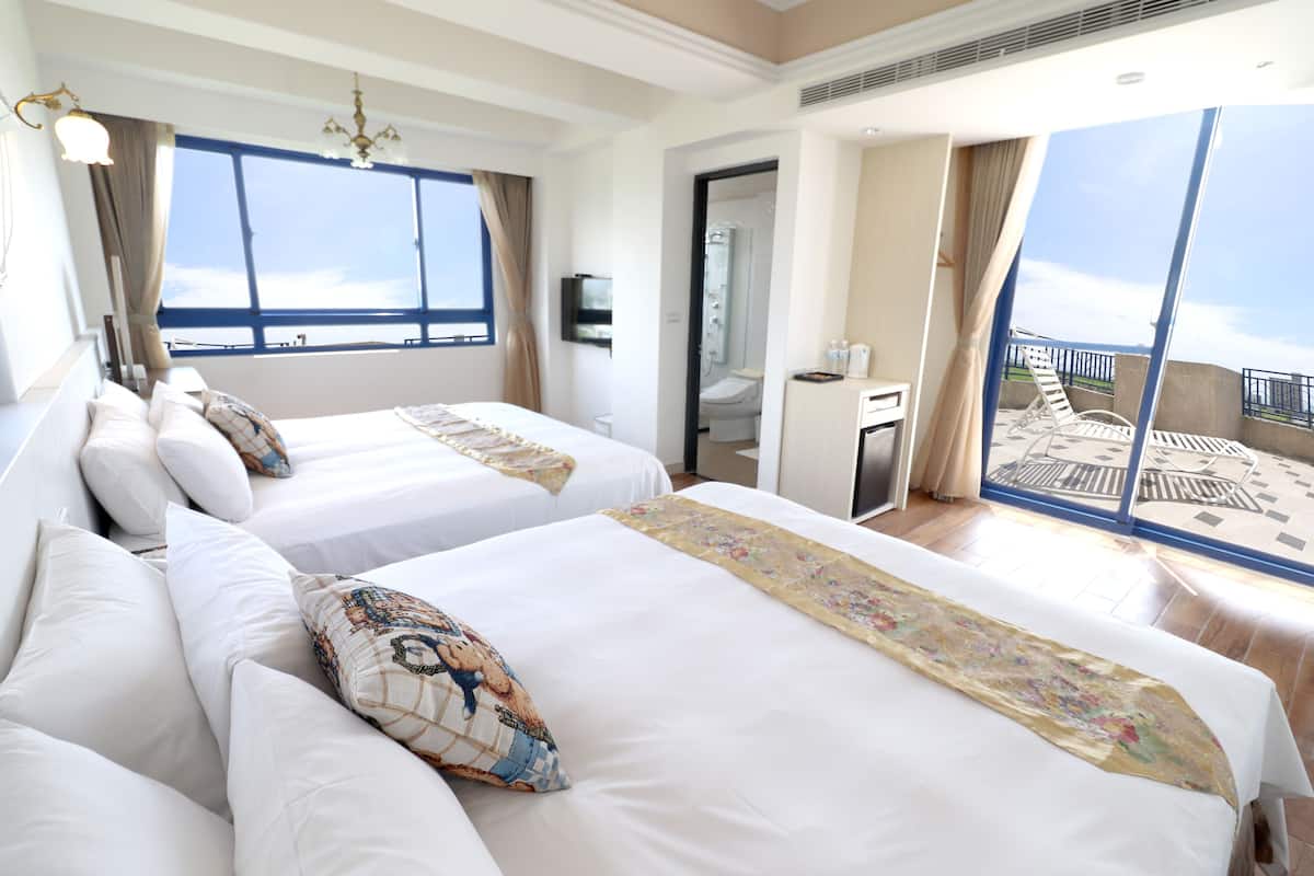 A hotel room with two beds, a large window, a balcony, and a bathroom visible through an open door.
