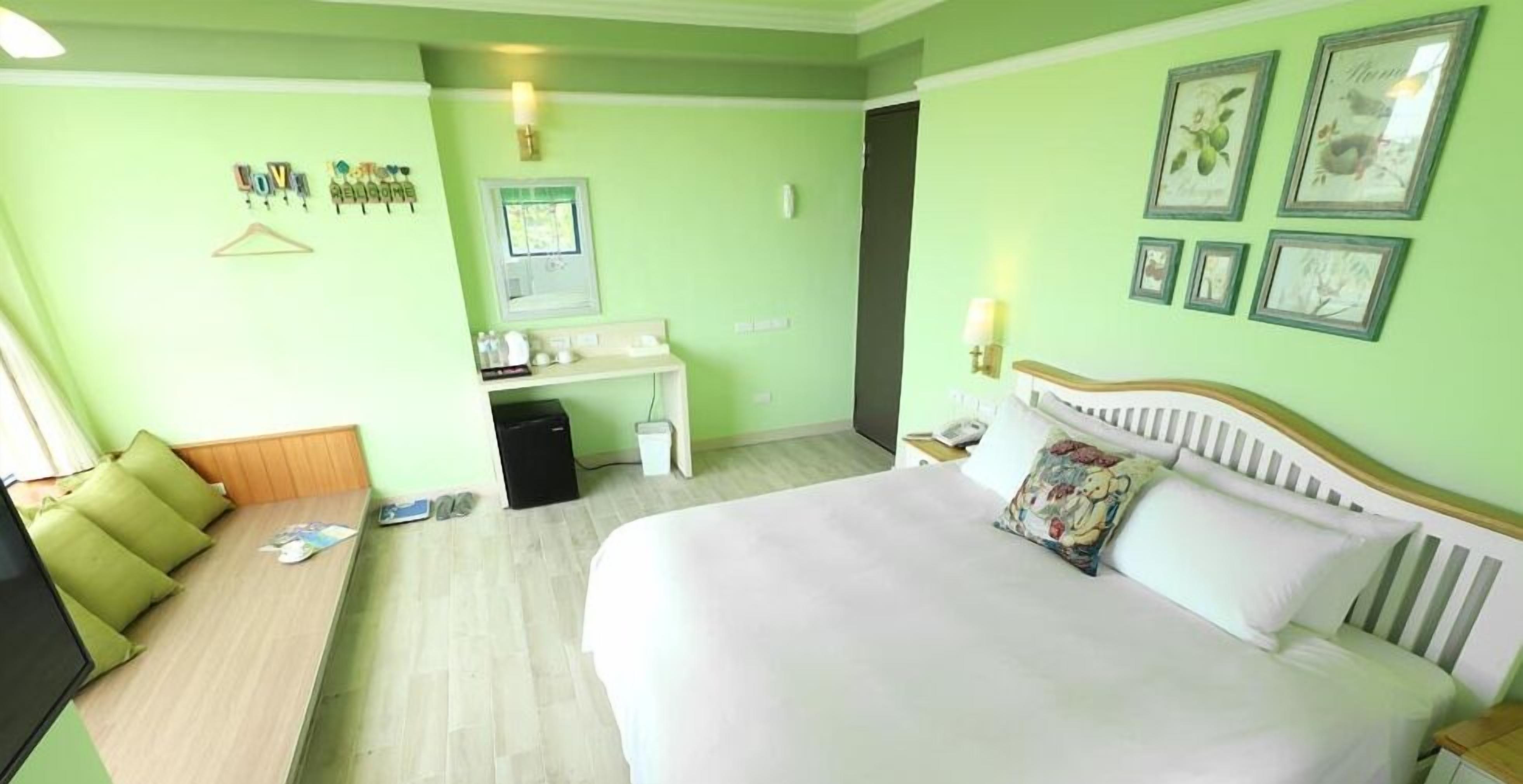A hotel room with a bed, a desk, a chair, a small table, a mirror, and a wall with framed pictures.