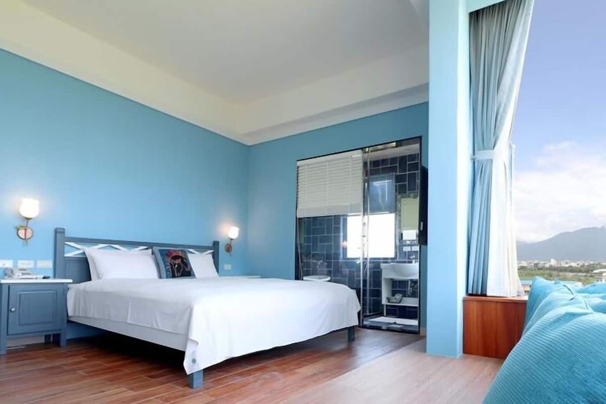 A bedroom with a bed, bedside tables, a bathroom with a glass shower, and a view of a mountain.