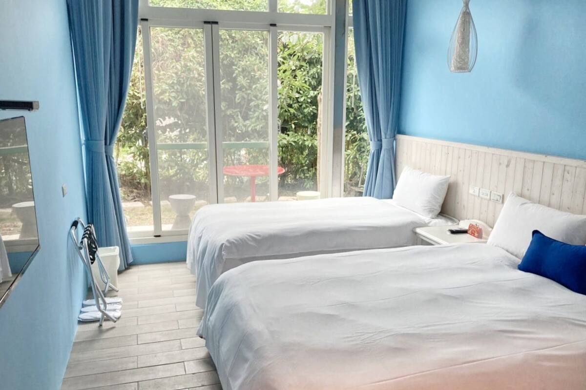 A bedroom with two beds, a window with curtains, a wooden floor, and a view of greenery outside.