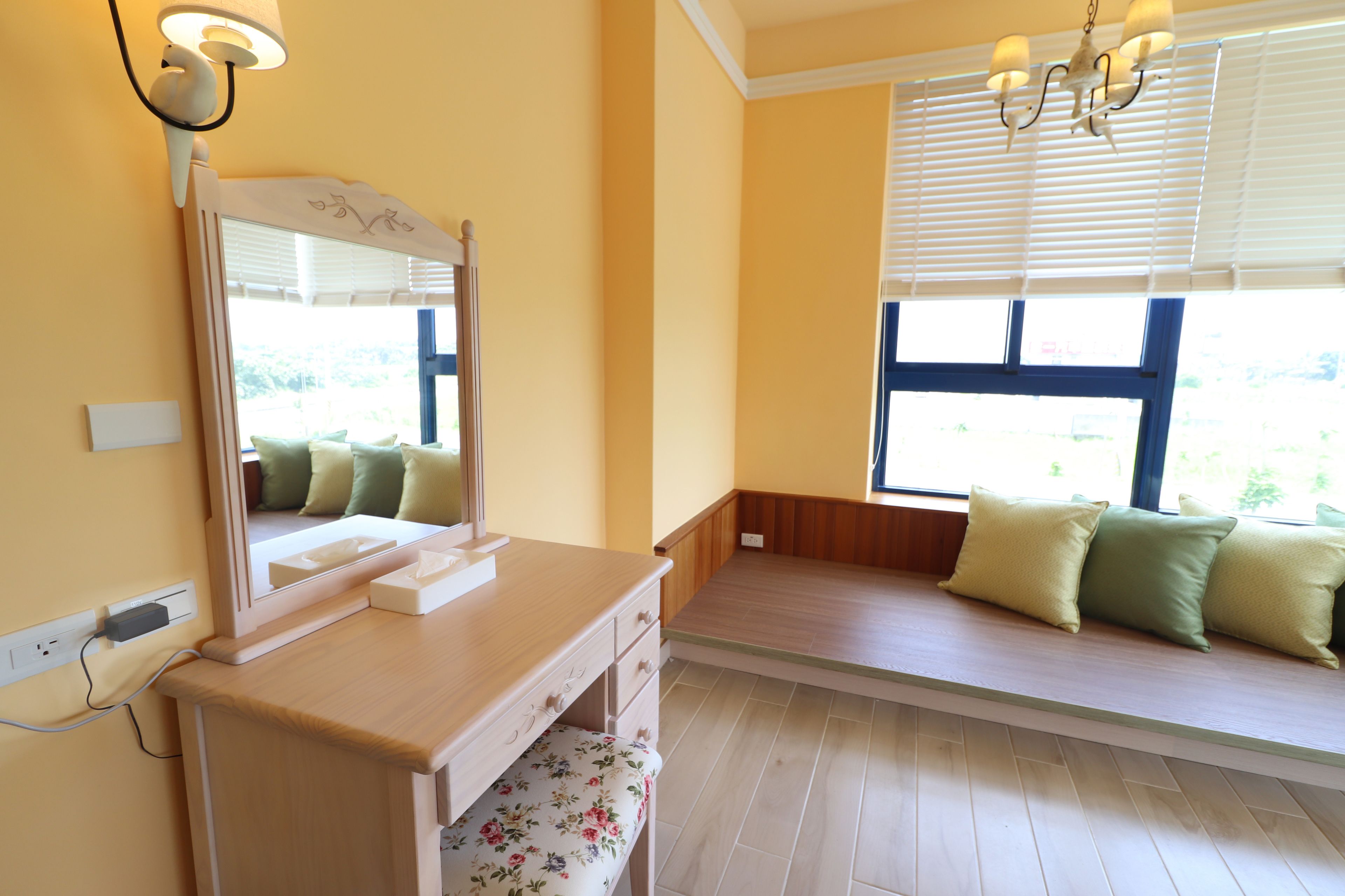 A room with a wooden desk, a mirror, a bench with cushions, and a window with blinds.