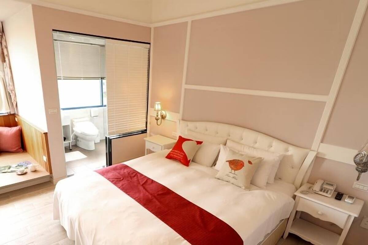 A hotel room with a large bed, a seating area with cushions, a bathroom with a toilet and sink, and a window with curtains.