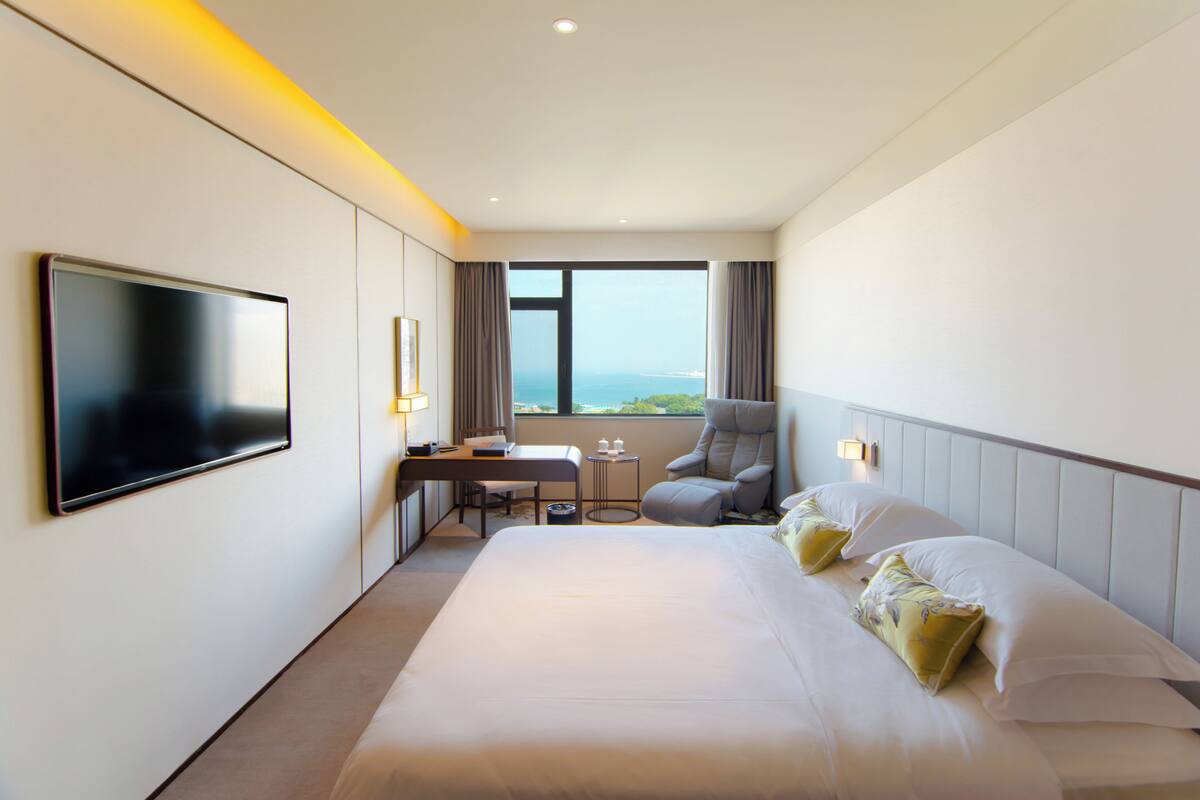 A hotel room with a large bed, a desk, a chair, a TV, and a view of the sea.