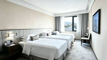 A hotel room with two beds, a desk, a chair, a nightstand, and a large window with a city view.