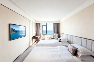 A hotel room with two beds, a desk, a chair, a TV, and a window with a view of the sea.