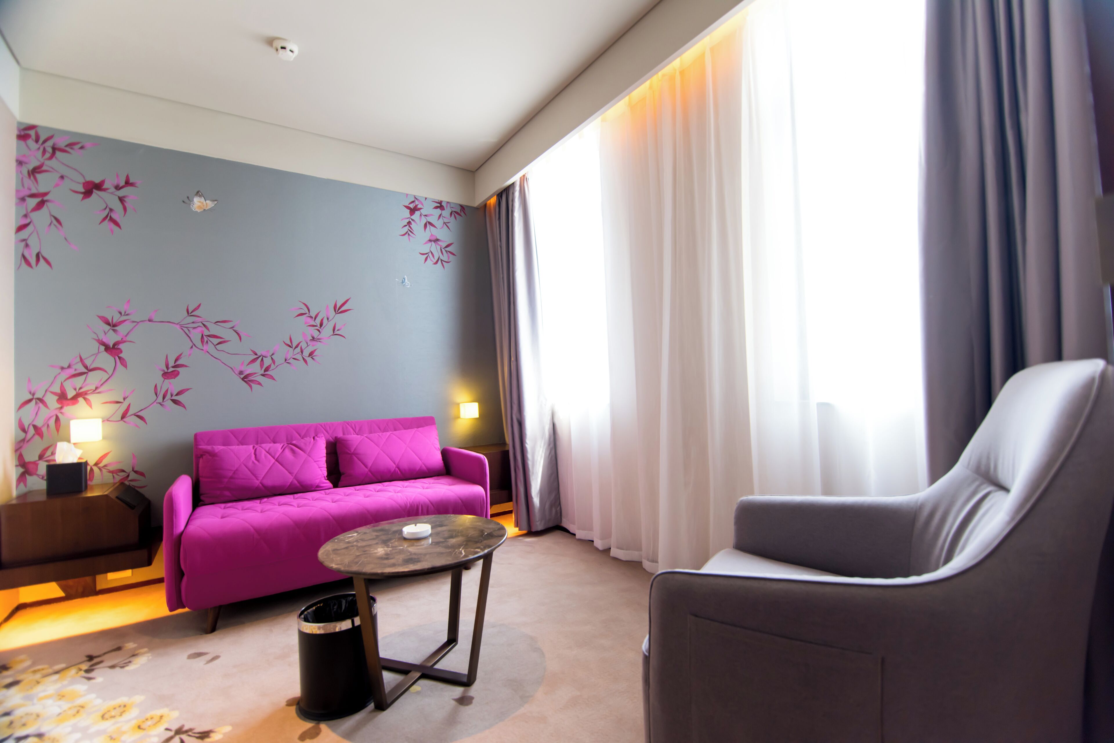 Una habitación de hotel moderna con un sofá púrpura, un sillón gris, una mesita redonda y una pared decorativa con motivos florales en rosa.