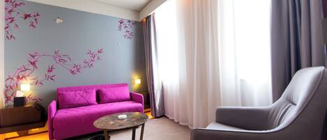 Una habitación de hotel moderna con un sofá púrpura, un sillón gris, una mesita redonda y una pared decorativa con motivos florales en rosa.