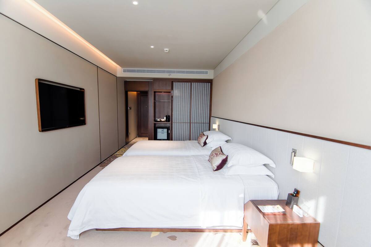 A hotel room with two beds, a TV mounted on the wall, a bedside table with a lamp, and a sliding door closet.