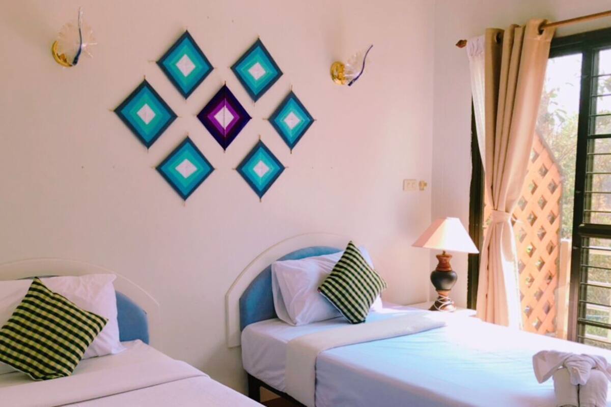 Quarto com duas camas, uma obra de arte na parede, uma luminária e uma janela com cortinas.