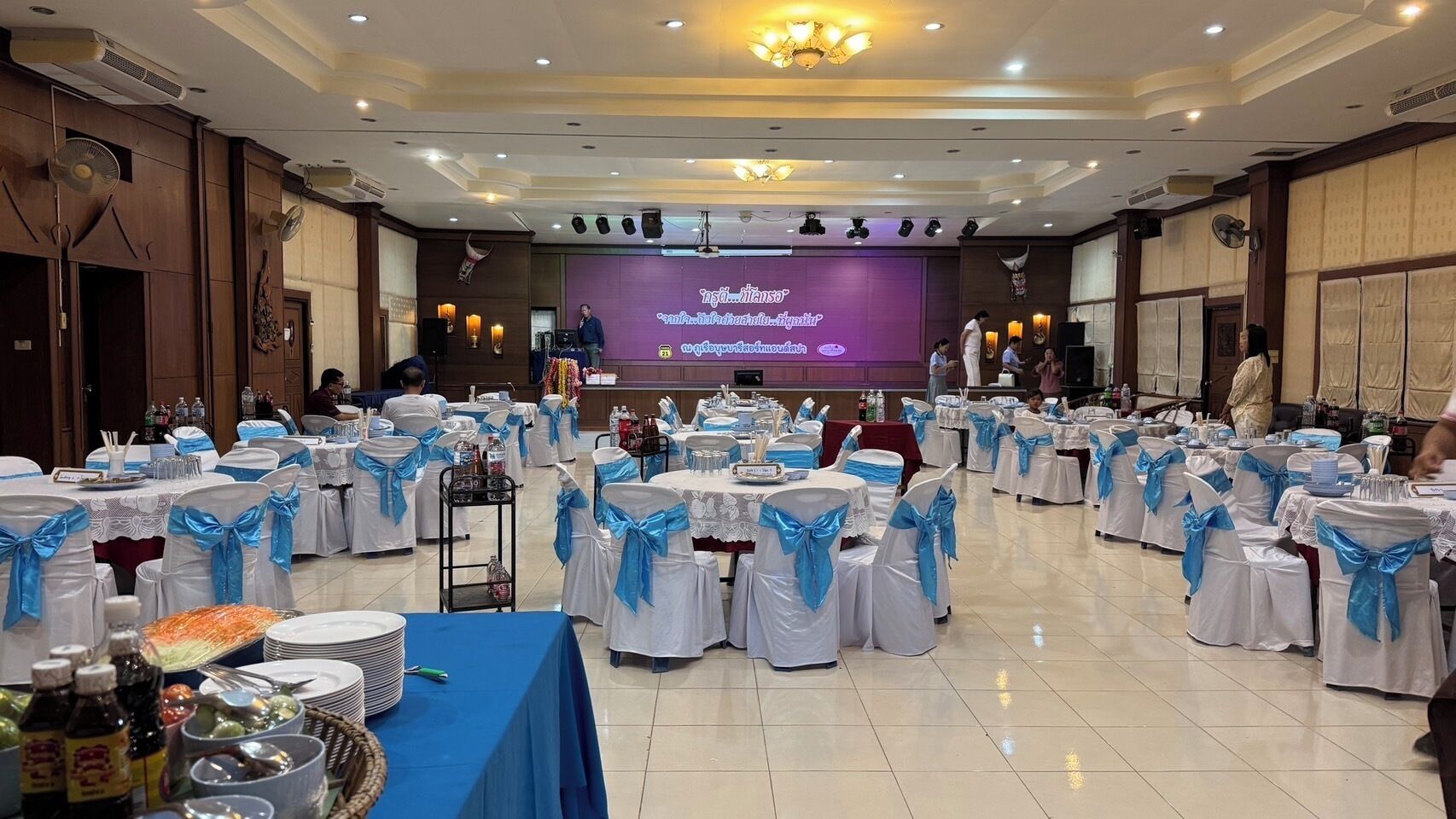 Un salón de banquetes con mesas redondas dispuestas para un evento formal, con cintas azules en las sillas y un escenario con una pantalla de presentación.