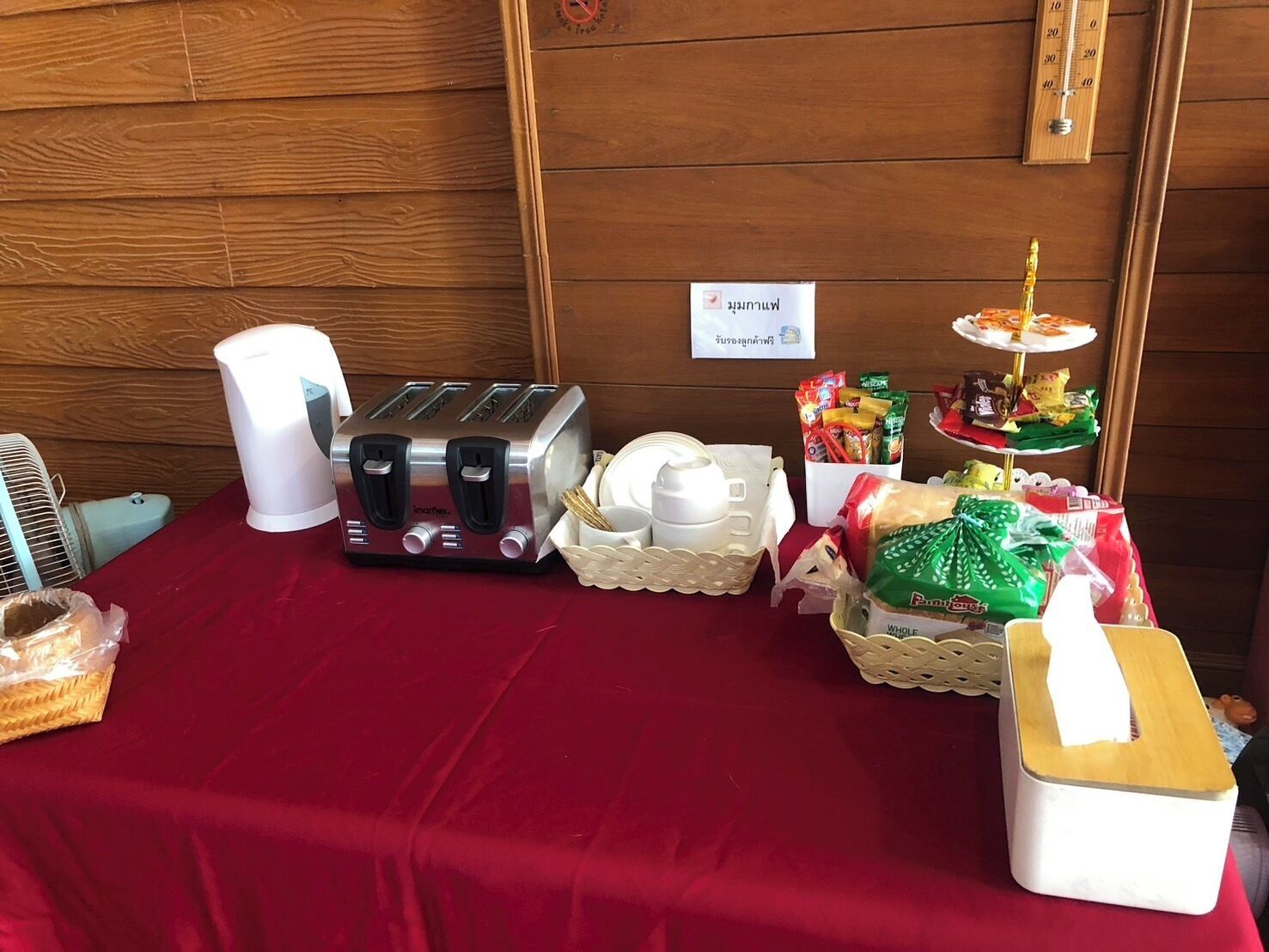 Una mesa con mantel rojo, un tostador, una jarra blanca, una cesta con tazas blancas, una bandeja con aperitivos y un pequeño soporte con una taza de café.