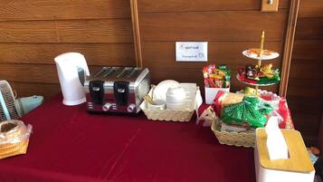 Una mesa con mantel rojo, un tostador, una jarra blanca, una cesta con tazas blancas, una bandeja con aperitivos y un pequeño soporte con una taza de café.