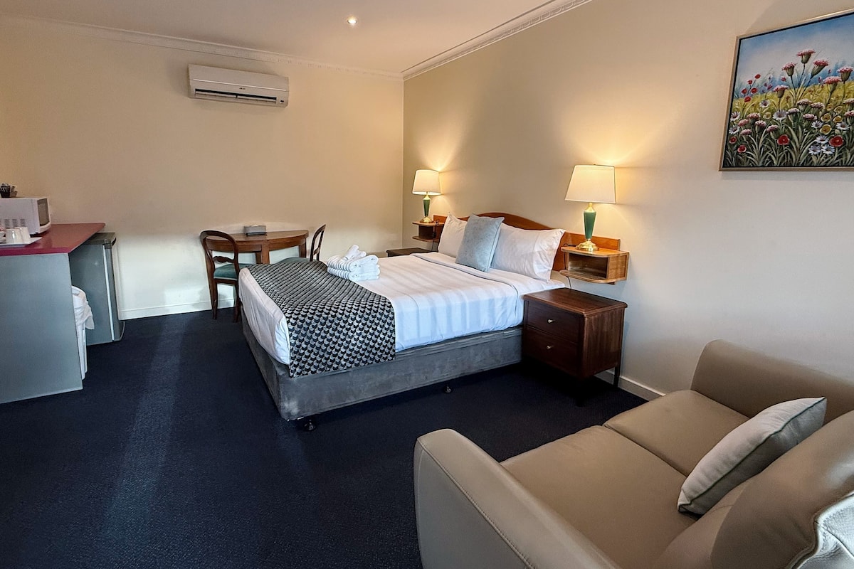 A hotel room with a bed, bedside tables, a desk, a chair, a couch, and a painting on the wall.